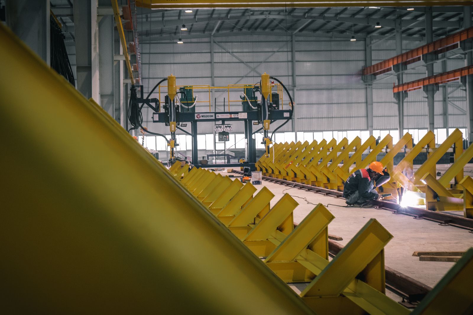 Lasser met oranje helm aan het werk tussen gele constructies in een grote werkhal